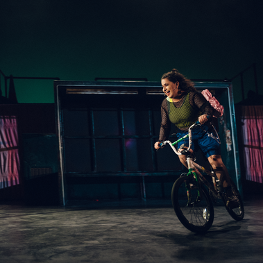 An individual is riding a BMX while smiling. They're dressed in odd and non-matching clothes, and there's a bus stop and a swing set behind them.