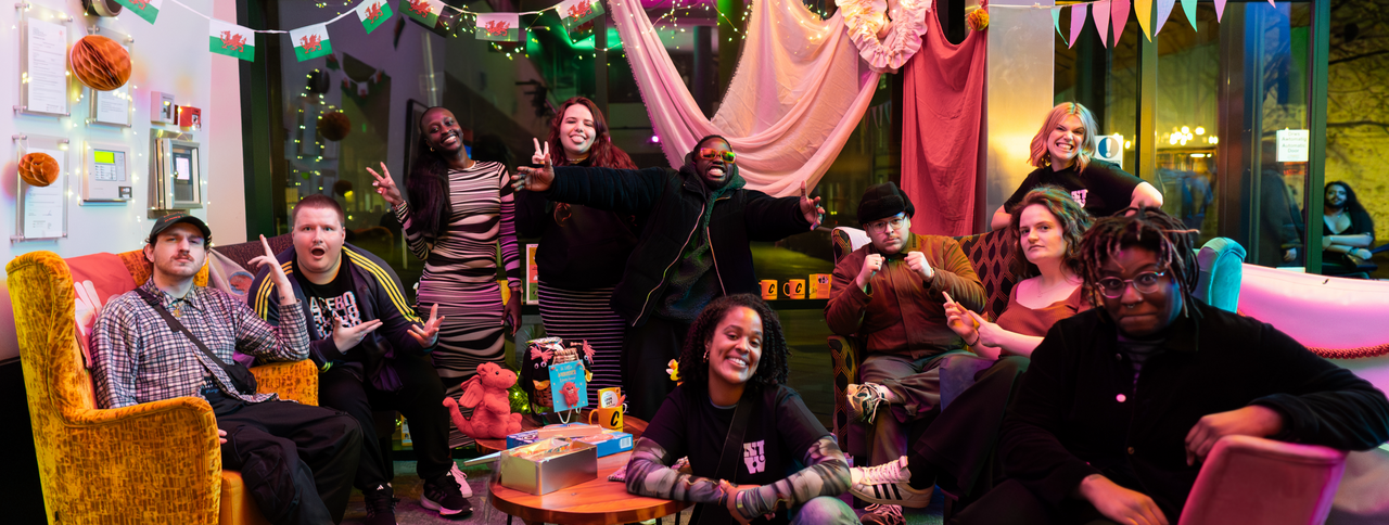 Ten individuals are gathered in a lounge area. They're all smiling at the camera and making different gestures with their hands.