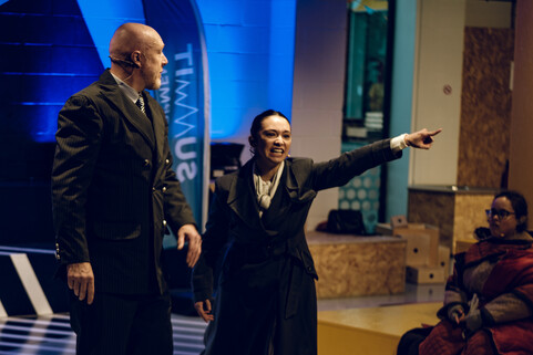 A man and woman dressed in suits. The woman is pointing into the distance angrily.