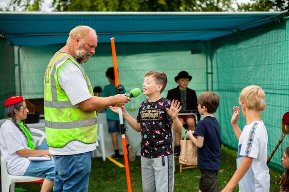 A man in a fluorescent vest holds a mic to a young boy with a lightsaber, there are other children in the background.