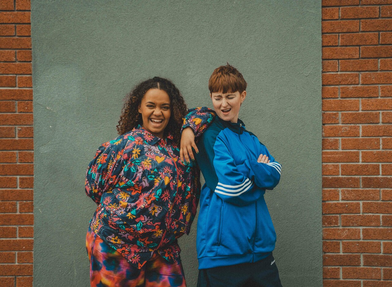 Two individuals are standing against a brick wall. The individual on the left has their arm on the other's shoulder and is smiling at the camera. The individual on the right is looking back at the other individual.