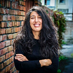 An individual with long black wavy hair. Their arms are crossed and they're looking at the camera.