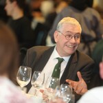 Unigolyn gyda gwallt llwyd byr a sbectol. Maen nhw wrth fwrdd gyda diodydd ac yn gwenu.