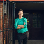An individual is standing, leaning against a gate. They have short brown hair and are wearing a green jumper.