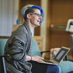 An individual with blonde and blue hair. They're wearing glasses and are sitting down with a laptop on their lap.