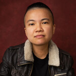 An individual with short brown hair and a brown jacket with a white collar. They're looking directly at the camera.