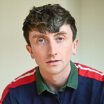 Unigolyn gyda gwallt brown byr, yn gwisgo crys polo glas, gwyrdd a choch ac yn edrych ar y camera.