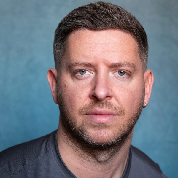 An individual with short brown hair and facial hair, wearing a grey shirt and looking at the camera.
