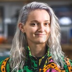 Unigolyn â gwallt gwyn hir, sy'n gorchuddio ochr chwith ei wyneb ac yn cuddio y tu ôl i'w glust dde. Maen nhw'n gwisgo siwmper liwgar ac yn gwenu ar y camera.