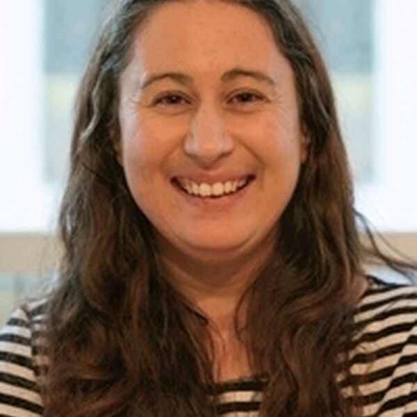 Unigolyn gyda gwallt hir brown a thop streipïog du a gwyn, yn gwenu ar y camera.