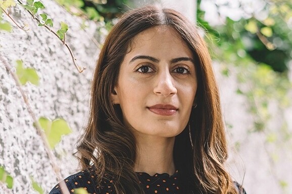 A woman with long brown hair looking directly into the camera, there are trees behind her.