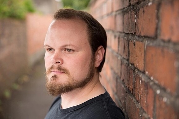 A man with a moustache and beard, with his back against a brick wall.