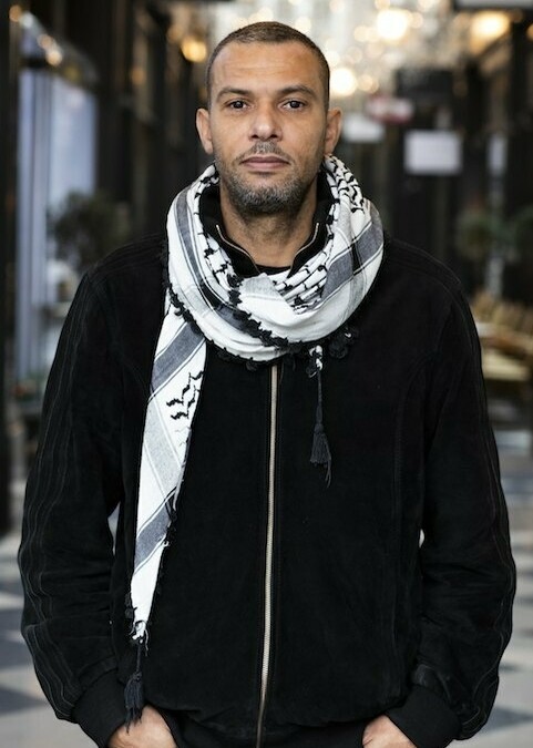Kyle is wearing a black jacket with a black and white scarf around his neck