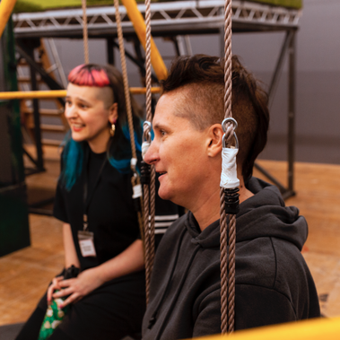 Two people are sat on swings. They're both looking ahead and smiling. One has a brown mohawk and is wearing a dark grey hoodie. The other has long pink and blue hair and is wearing a black t-shirt.