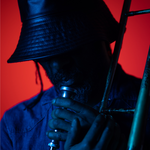 Llun pen-ymlaen o unigolyn sy'n edrych i lawr. Maen nhw’n gwisgo het fwced sy’n gorchuddio eu llygaid ac maen nhw’n dal trombone. Mae'r golau yn las a choch.