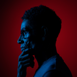 An individual’s profile as they’re looking to the left. They are wearing glasses and their hand is on their chin. The lighting is blue and red.