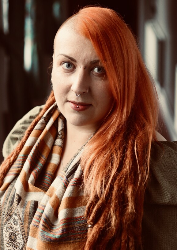An individual with long red hair. They have a nose and lip piercing and are looking at the camera.